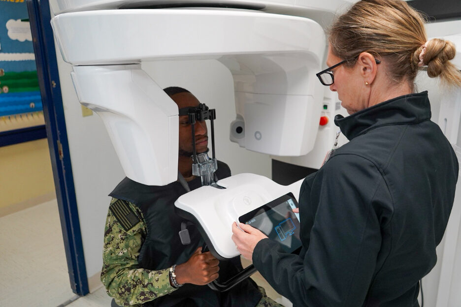 Patient positioned in a cone beam CT scanner while a clinician reviews the scan on a tablet control panel