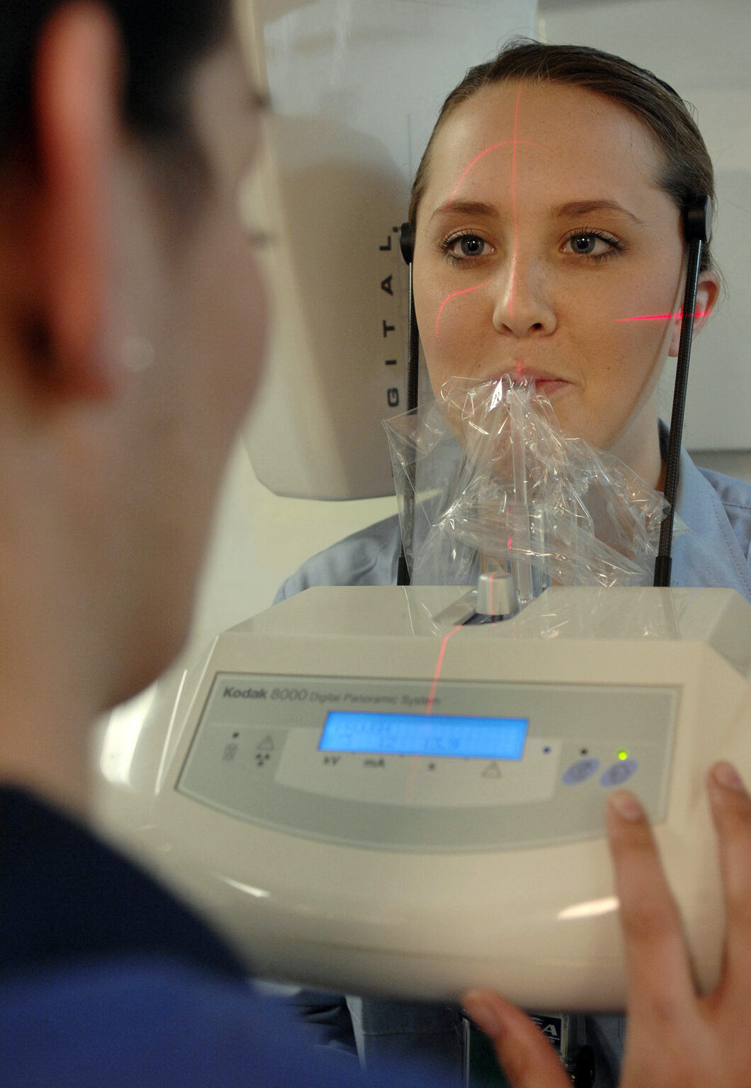 Patient positioned in a digital panoramic dental X-ray system while a technician operates the controls